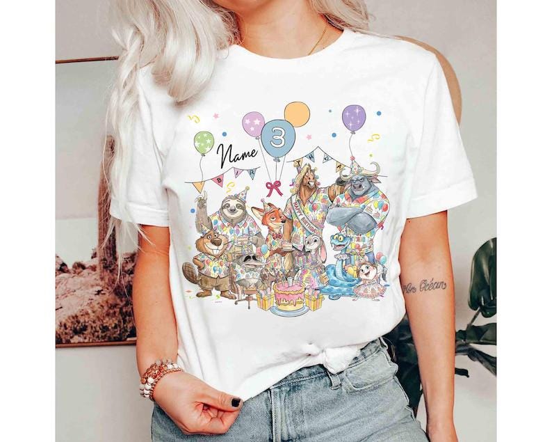 May include: White t-shirt featuring a colorful birthday party scene with cartoon animals, balloons, and the number "3". The word "Name" is written above the characters. The design includes a cake and festive decorations.