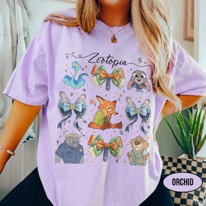 May include: A light purple t-shirt featuring the text Zootopia and a graphic print of various characters including a fox, a rabbit, and a buffalo, interspersed with several decorative bows and small star shapes across the front of the garment.