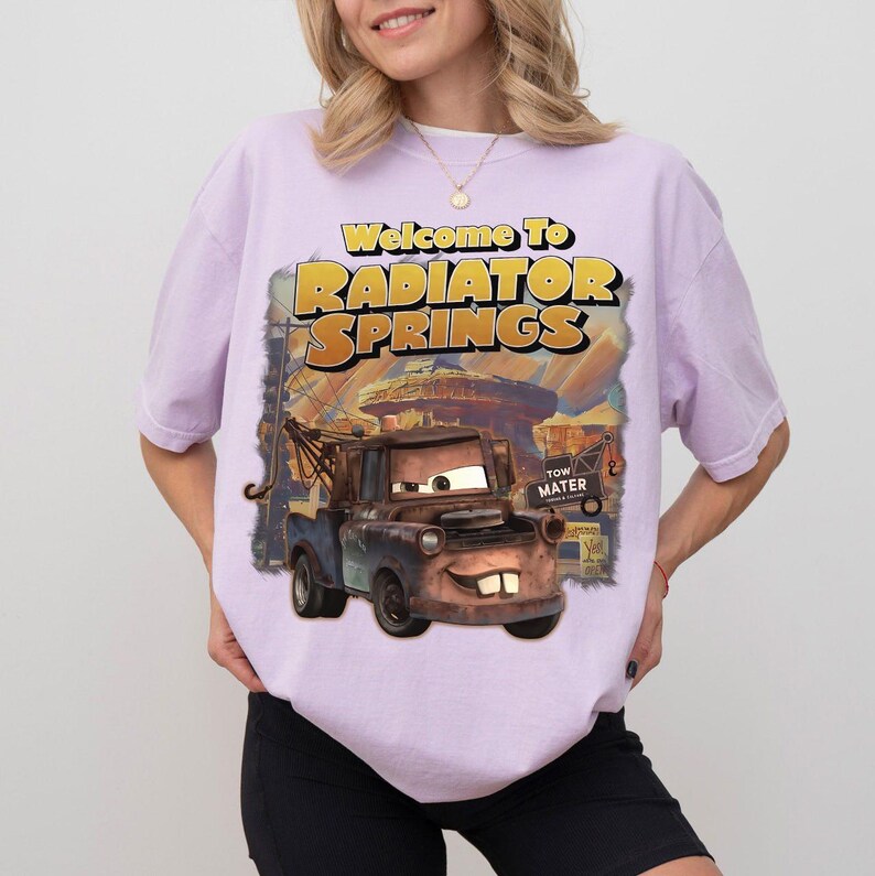 May include: A light purple t-shirt featuring a graphic print of the character Mater from the movie Cars. The text reads Welcome To RADIATOR SPRINGS and TOW MATER. The design shows a rusty brown tow truck with large eyes and prominent front teeth.