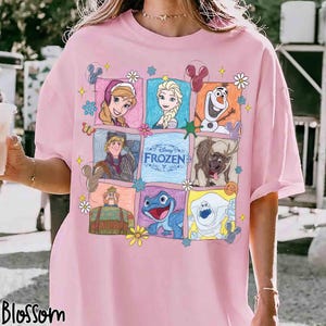 May include: Pale pink t-shirt with a colourful cartoon grid of characters from Disney's "Frozen." The grid includes Anna, Elsa, Olaf, Kristoff, Sven, and other characters, with the word "Frozen" in the centre. The word "Blossom" is at the bottom left.