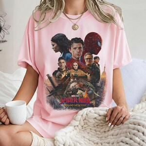 May include: A light pink t-shirt featuring a graphic from the movie "Spider-Man: Brand New Day." The design includes several characters, the Spider-Man mask, and the movie title. The shirt is a crew neck style.
