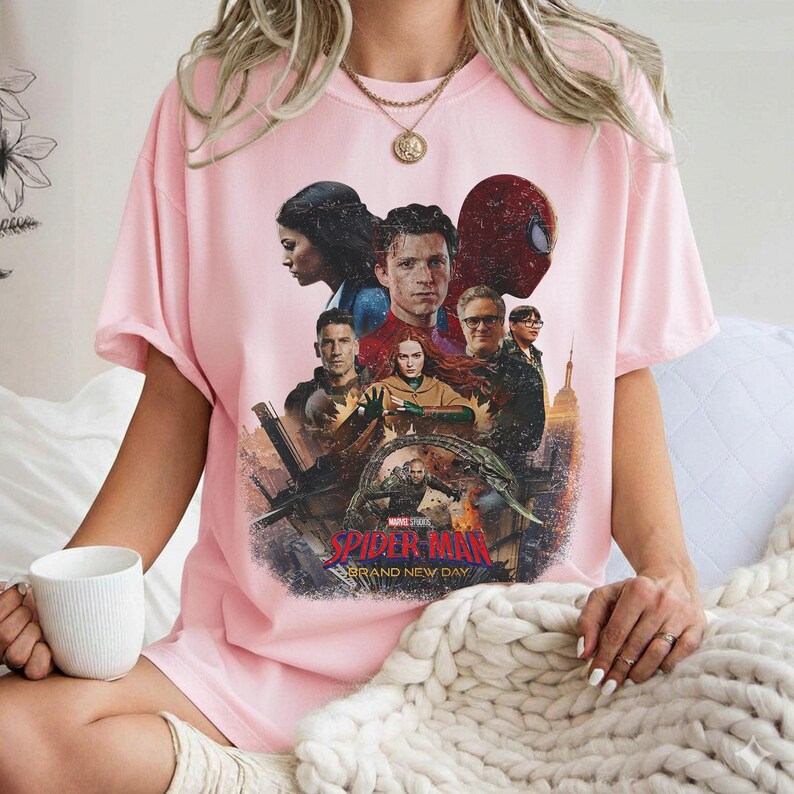 May include: A light pink t-shirt featuring a graphic from the movie "Spider-Man: Brand New Day." The design includes several characters, the Spider-Man mask, and the movie title. The shirt is a crew neck style.
