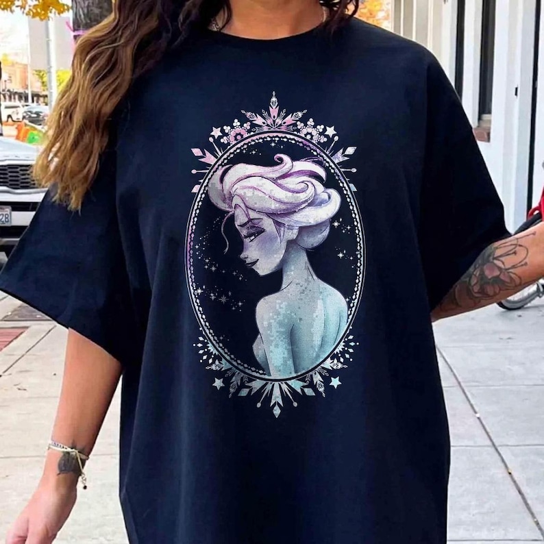 May include: A navy blue t-shirt featuring a detailed illustration of a woman with white hair and a blue-toned complexion, set within an oval frame. The frame is adorned with a decorative border of snowflakes and crystals.