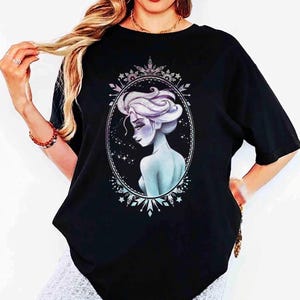 May include: Black t-shirt featuring a detailed illustration of a woman with light blue skin and white hair, framed within an oval border. The design has a celestial theme with stars and snowflake accents.