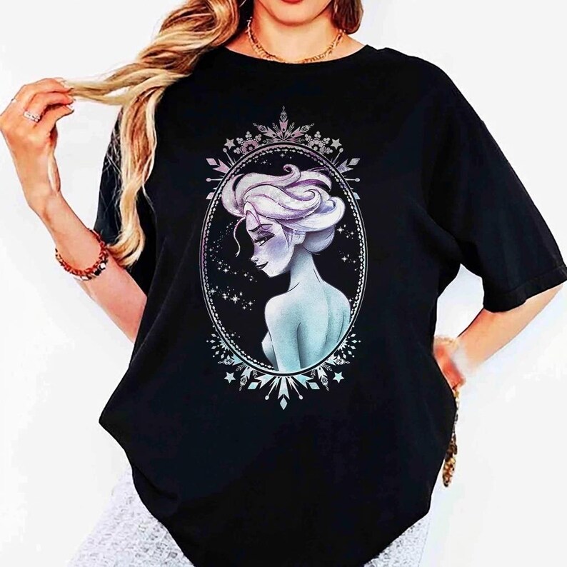 May include: Black t-shirt featuring a detailed illustration of a woman with light blue skin and white hair, framed within an oval border. The design has a celestial theme with stars and snowflake accents.