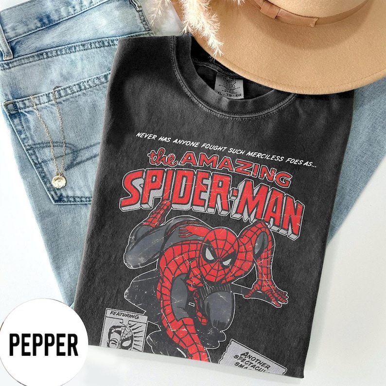 May include: A black t-shirt featuring a vintage Spider-Man graphic. The design includes the words "the AMAZING SPIDER-MAN" in red and white, with an illustration of Spider-Man. The shirt is paired with light blue jeans and a tan hat.