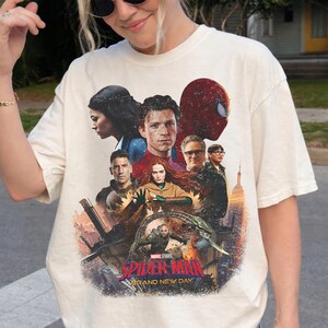 May include: Cream-colored t-shirt featuring a graphic of characters from the Marvel Studios film "Spider-Man: Brand New Day." The design includes images of Spider-Man, Tom Holland, and other actors. The text "Spider-Man" and "Brand New Day" are also visible.