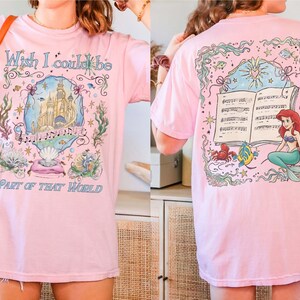 May include: Pink t-shirt featuring a colorful illustration from "The Little Mermaid." The front design includes a castle, musical notes, and text that reads "Wish I could be part of that world." The back shows Ariel, a crab, and a fish.