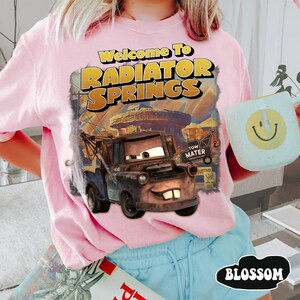 May include: A light pink t-shirt featuring a graphic print of the character Tow Mater from the movie Cars. The text reads Welcome To RADIATOR SPRINGS and TOW MATER. The design includes a desert landscape background with rock formations.