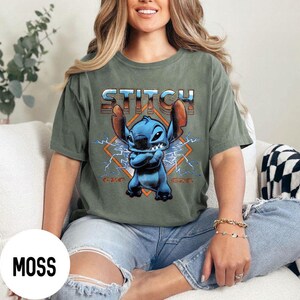 May include: Moss green t-shirt featuring a blue cartoon character with large ears and the word "STITCH" in a retro font. The character is set against a diamond-shaped background with lightning bolts.