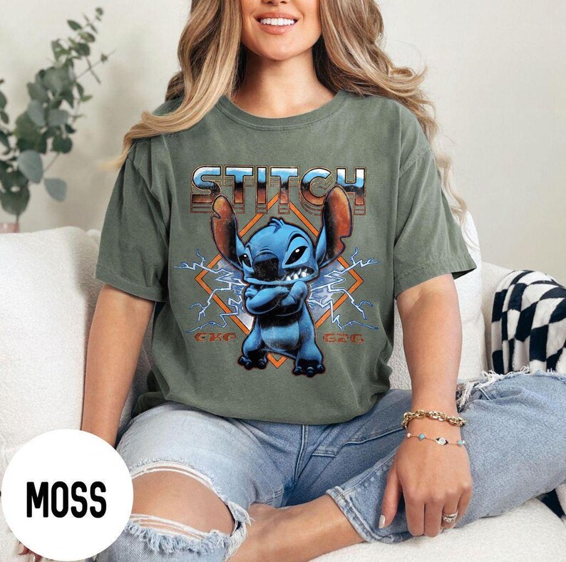 May include: Moss green t-shirt featuring a blue cartoon character with large ears and the word "STITCH" in a retro font. The character is set against a diamond-shaped background with lightning bolts.
