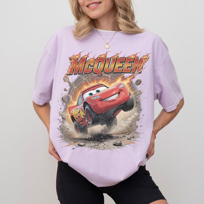May include: A light purple t-shirt featuring a graphic of Lightning McQueen from the movie Cars. The red race car is jumping with the word "McQueen" above it in a fiery font. The shirt is a casual, oversized fit.