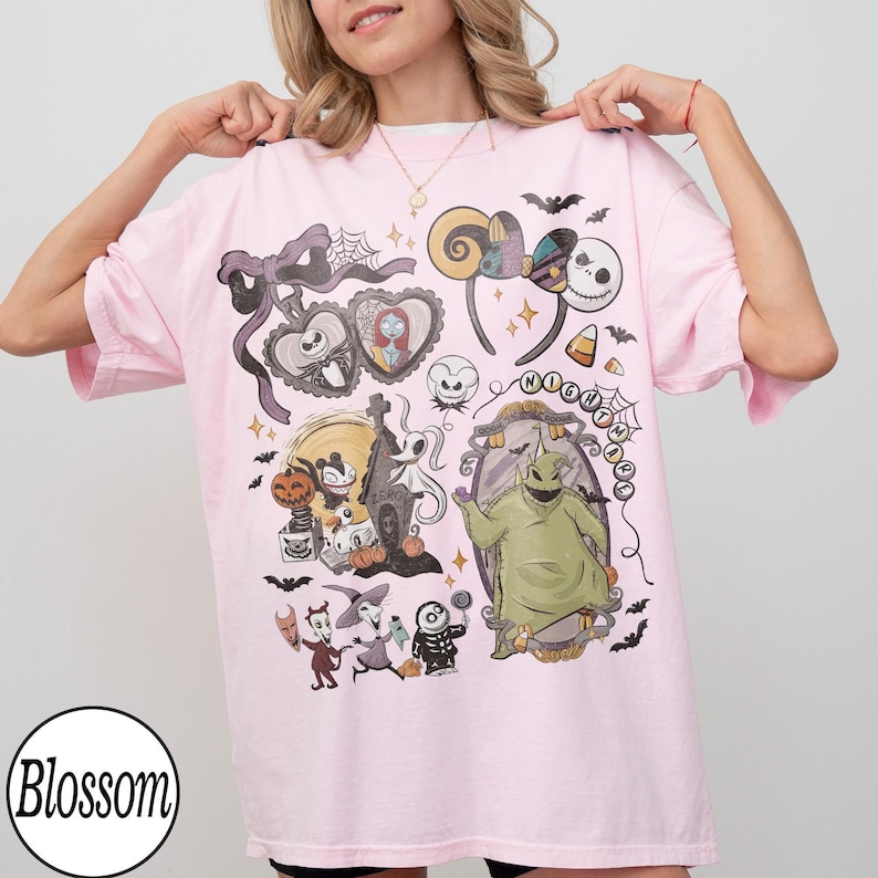 May include: A light pink t-shirt featuring a graphic design with characters and elements from "The Nightmare Before Christmas." The design includes Jack Skellington, Sally, Oogie Boogie, and other Halloween-themed imagery. The shirt has a round neckline and short sleeves.