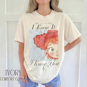 May include: An ivory short-sleeve t-shirt featuring a graphic print of a cartoon character wearing a red cowboy hat. The text reads EST 2026, I Knew It, and I Knew You. The design has a watercolor aesthetic with a light blue square background.