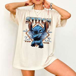 May include: Cream-colored t-shirt featuring a blue cartoon character with the word "STITCH" above it. The character is in a diamond-shaped design with lightning bolts. The text "G26" and "EKP" are also visible.