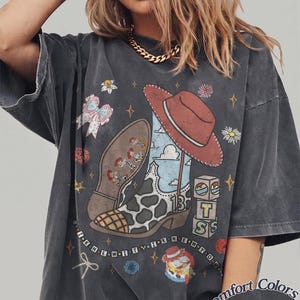 May include: A charcoal grey short-sleeve t-shirt featuring a graphic print of a cowboy boot with a cow-print pattern, a red hat, building blocks, and small character figures. The text "I KNEW IT I KNEW YOU" is printed in a line across the lower graphic area.