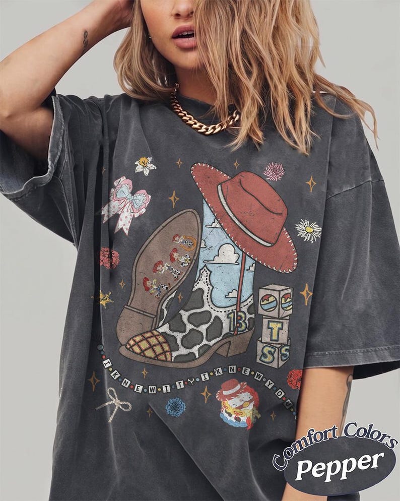 May include: A charcoal grey short-sleeve t-shirt featuring a graphic print of a cowboy boot with a cow-print pattern, a red hat, building blocks, and small character figures. The text "I KNEW IT I KNEW YOU" is printed in a line across the lower graphic area.