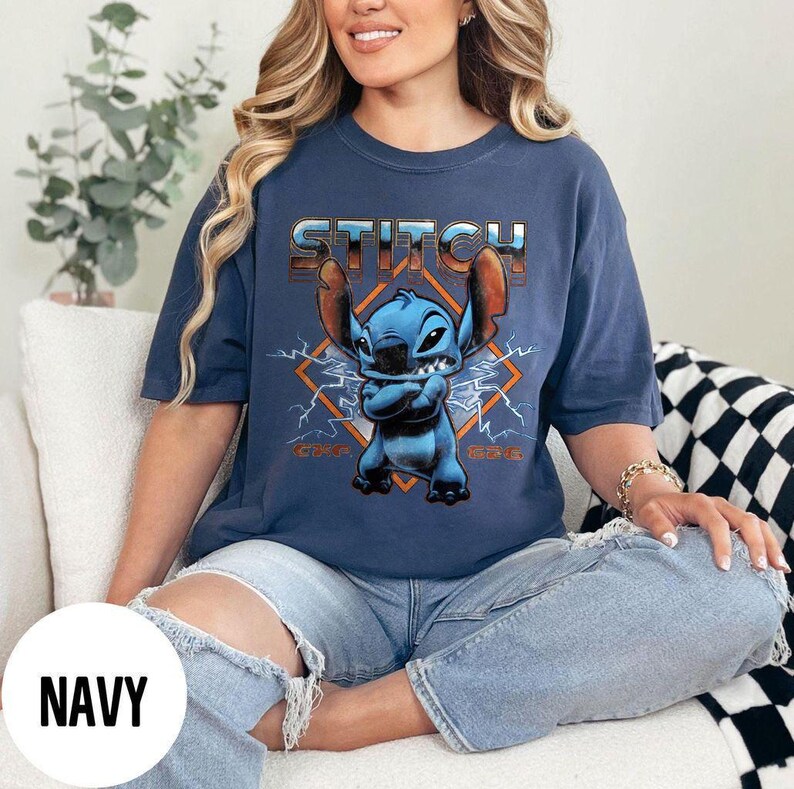 May include: Navy blue t-shirt featuring the character Stitch from Lilo & Stitch. The design includes the word "Stitch" in a metallic font above an image of Stitch with orange lightning bolts and a diamond shape.