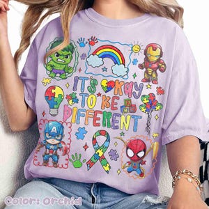 K&ouml;nnte beinhalten: Orchideenfarbenes T-Shirt mit bunten Cartoon-Illustrationen von Superhelden und einem Regenbogen. Das Shirt tr&auml;gt den Text "It's Okay to Be Different" in einer verspielten Schriftart, zusammen mit Puzzleteilen und Handabdr&uuml;cken.