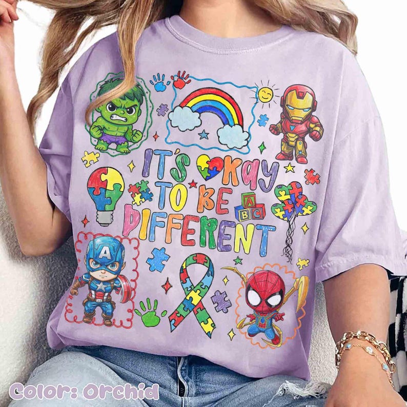 K&ouml;nnte beinhalten: Orchideenfarbenes T-Shirt mit bunten Cartoon-Illustrationen von Superhelden und einem Regenbogen. Das Shirt tr&auml;gt den Text "It's Okay to Be Different" in einer verspielten Schriftart, zusammen mit Puzzleteilen und Handabdr&uuml;cken.