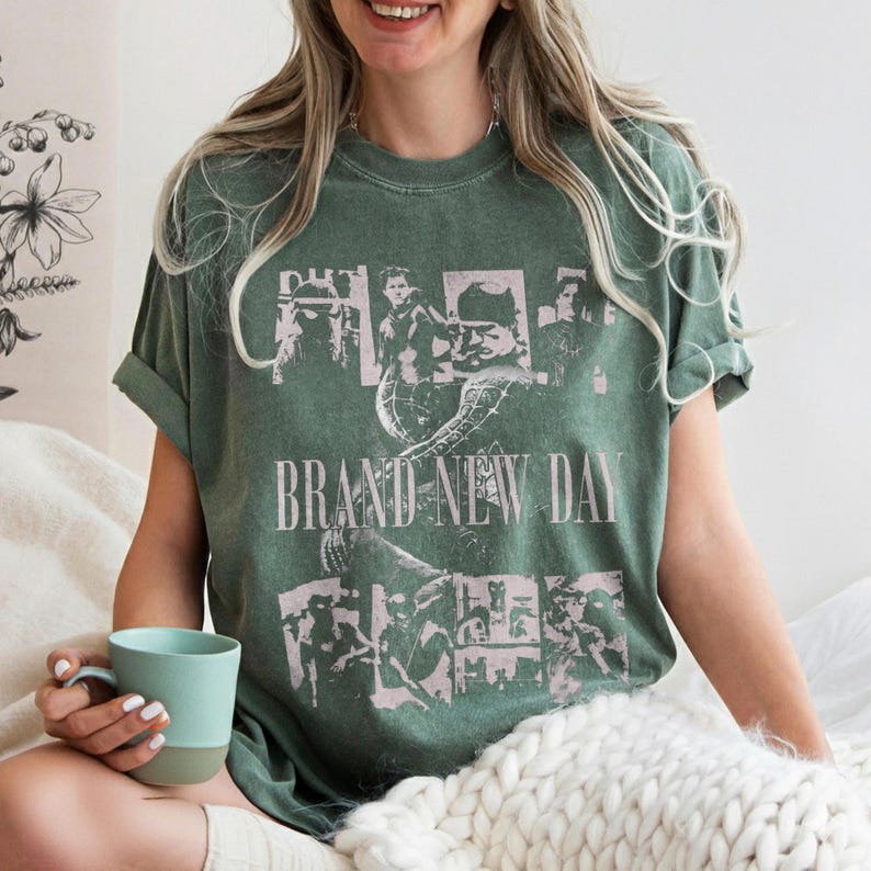 May include: Green t-shirt with a graphic design featuring multiple images and the text "BRAND NEW DAY" in a light pink color. The shirt has rolled-up sleeves and is worn with a light blue and gray mug.
