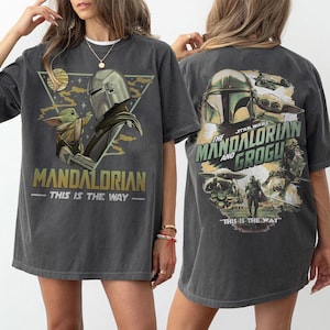 May include: Dark grey t-shirt with a graphic of The Mandalorian and Grogu. The front of the shirt has the text "MANDALORIAN - THIS IS THE WAY" and the back has "STAR WARS THE MANDALORIAN AND GROGU - THIS IS THE WAY".