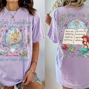 May include: A light purple t-shirt featuring a colorful illustration of a castle, musical notes, and seashells. The text "Wish I could be Part of That World" is displayed on the front. The back of the shirt shows a mermaid with a musical score.