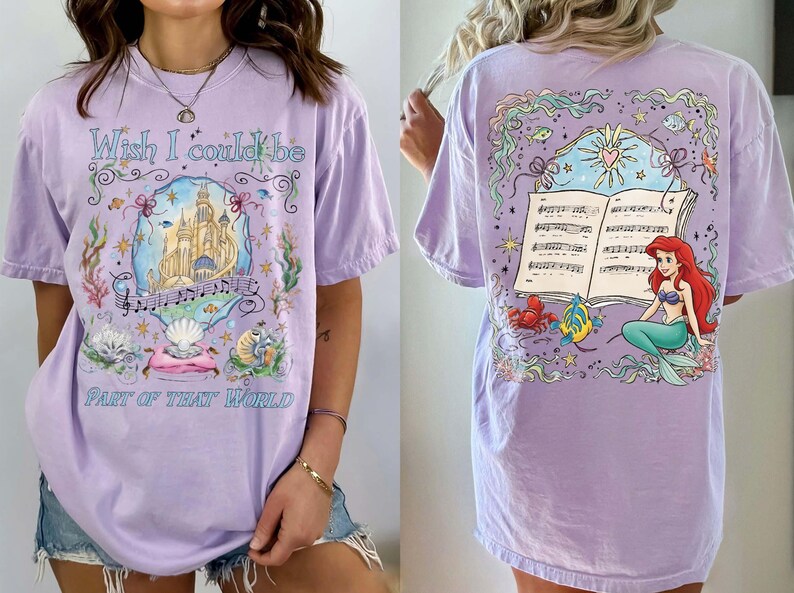 May include: A light purple t-shirt featuring a colorful illustration of a castle, musical notes, and seashells. The text "Wish I could be Part of That World" is displayed on the front. The back of the shirt shows a mermaid with a musical score.
