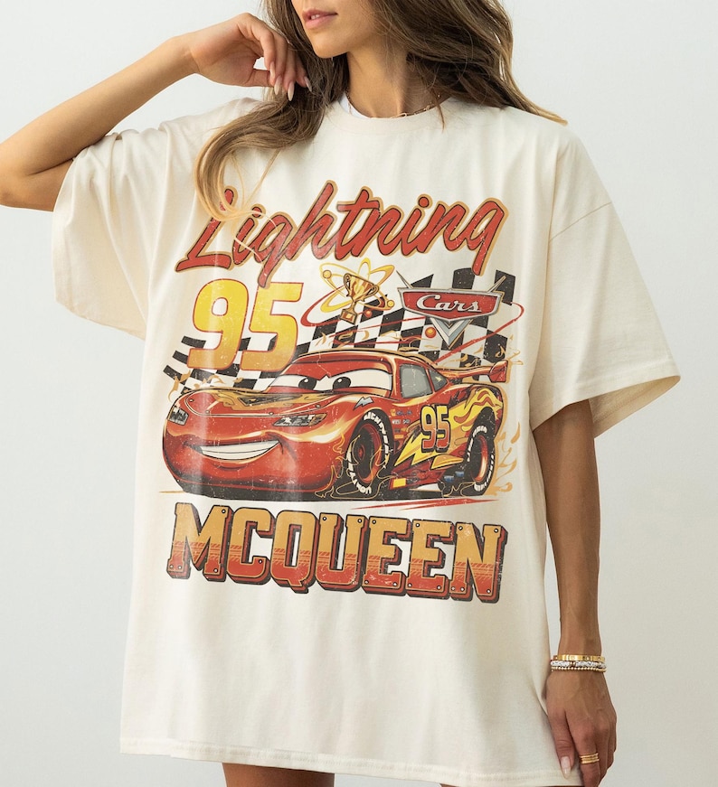May include: Cream-colored t-shirt featuring a graphic of Lightning McQueen from the Cars movie. The design includes the text "Lightning McQueen 95" in red and yellow, with a checkered flag and a trophy.