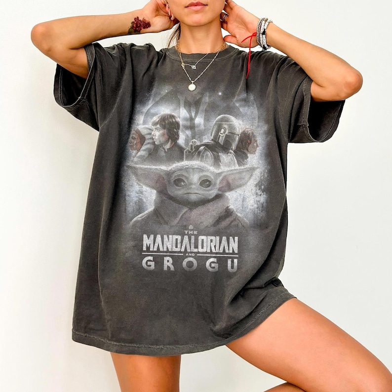 Op de afbeelding: Donkergrijs t-shirt met een afbeelding uit de serie "The Mandalorian". Het ontwerp toont Grogu en The Mandalorian, met de titel van de show eronder gedrukt. Het shirt heeft korte mouwen en een relaxte pasvorm.