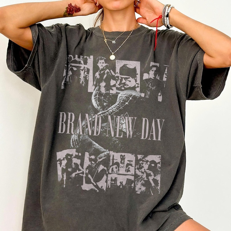 May include: Dark gray t-shirt with a graphic design featuring multiple black and white images and the text "BRAND NEW DAY" in large, white capital letters. The shirt has short sleeves and a relaxed fit.
