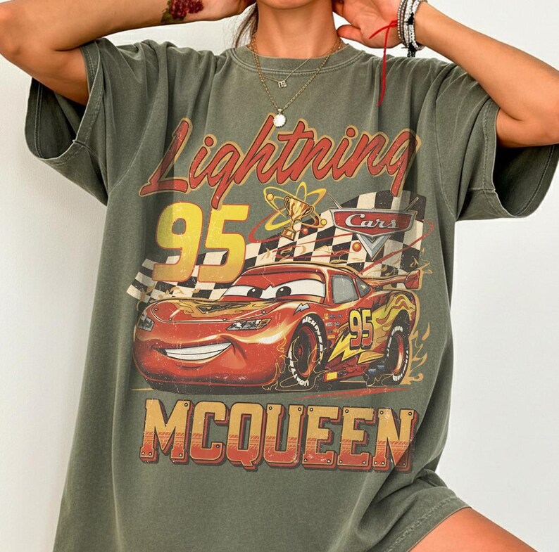 May include: Olive green t-shirt featuring a graphic of Lightning McQueen from the movie Cars. The design includes the text "Lightning 95" and "McQueen" in a vintage style. The shirt has short sleeves and a relaxed fit.