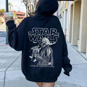 Może przedstawiać: Czarna bluza z kapturem z grafiką Star Wars na plecach. Projekt zawiera logo Star Wars, ilustrację The Mandalorian i Grogu oraz tekst "I'M A MANDALORIAN. WEAPONS ARE PART OF MY RELIGION."