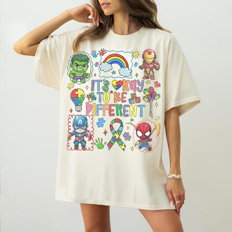 K&ouml;nnte beinhalten: Cremefarbenes T-Shirt mit farbenfrohen Cartoon-Illustrationen von Superhelden und dem Schriftzug "It's Okay to Be Different". Das Design beinhaltet einen Regenbogen, Puzzleteile und ein Band, das Inklusion und Akzeptanz f&ouml;rdert.