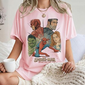 May include: A light pink t-shirt featuring a graphic of Spider-Man and other Marvel characters. The design includes the text "SPIDER-MAN BRAND NEW DAY". The shirt is a short-sleeved, casual style.