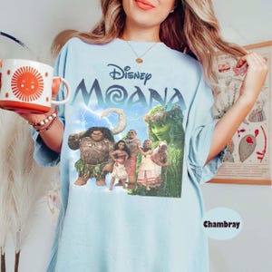 以下が含まれることがあります： ディズニー映画「モアナ」のロゴとキャラクターが描かれた水色のTシャツ。グラフィックには、マウイ、モアナ、その他のアニメのキャラクターが含まれています。太陽のデザインの白とオレンジのマグカップが画像に保持されています。