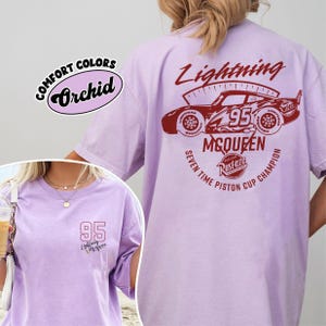 May include: A light purple t-shirt with a graphic of a race car and the text "Lightning McQueen 95". The shirt also features the text "Seven Time Piston Cup Champion". The shirt is a Comfort Colors brand.