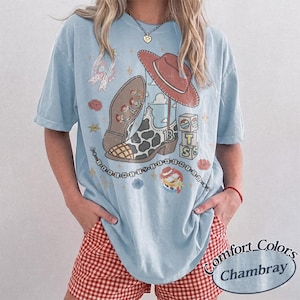 May include: A light blue short-sleeve t-shirt featuring a graphic print of a cow-print cowboy boot, a red hat, building blocks with the letters T, S, and a ball, and a string of beads with the text I KNEW IT I KNEW IT I KNEW IT.