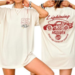 May include: Cream-colored t-shirt featuring a red graphic of Lightning McQueen and the number 95. The front has a small logo, while the back displays a larger design with text: "Seven Time Piston Cup Champion."