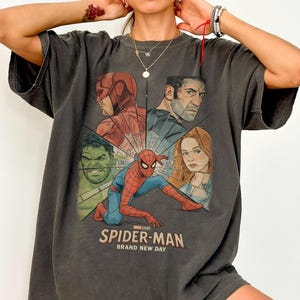 May include: Dark gray t-shirt featuring a graphic of Spider-Man and other Marvel characters. The design includes the text "SPIDER-MAN BRAND NEW DAY". The shirt has short sleeves and a crew neck.