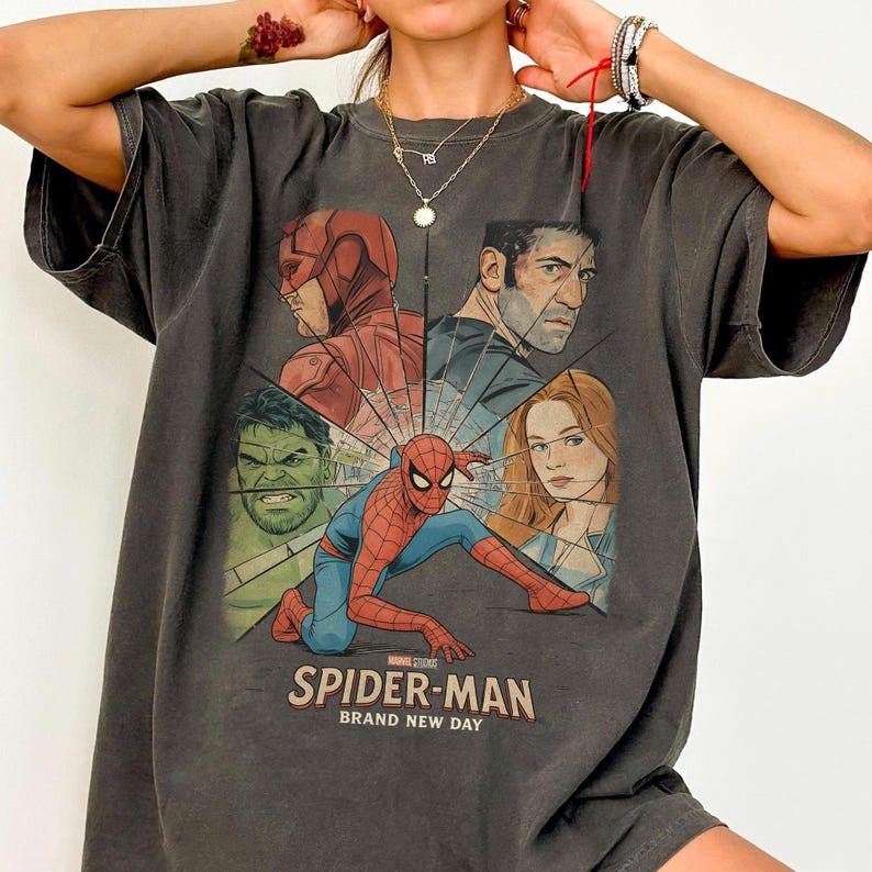 May include: Dark gray t-shirt featuring a graphic of Spider-Man and other Marvel characters. The design includes the text "SPIDER-MAN BRAND NEW DAY". The shirt has short sleeves and a crew neck.