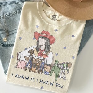 以下が含まれることがあります： 赤いカウボーイハットをかぶった人物と、その周りに描かれた様々なキャラクターのおもちゃや青い星が特徴のクリーム色のTシャツ。グラフィックの下部には青い文字で「I KNEW IT, I KNEW YOU」とプリントされています。
