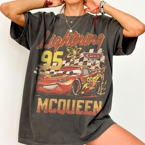 May include: Dark gray t-shirt featuring a graphic of Lightning McQueen from the Cars movie. The design includes the text "Lightning 95 McQueen" in yellow and red, with a checkered flag and atomic symbol.