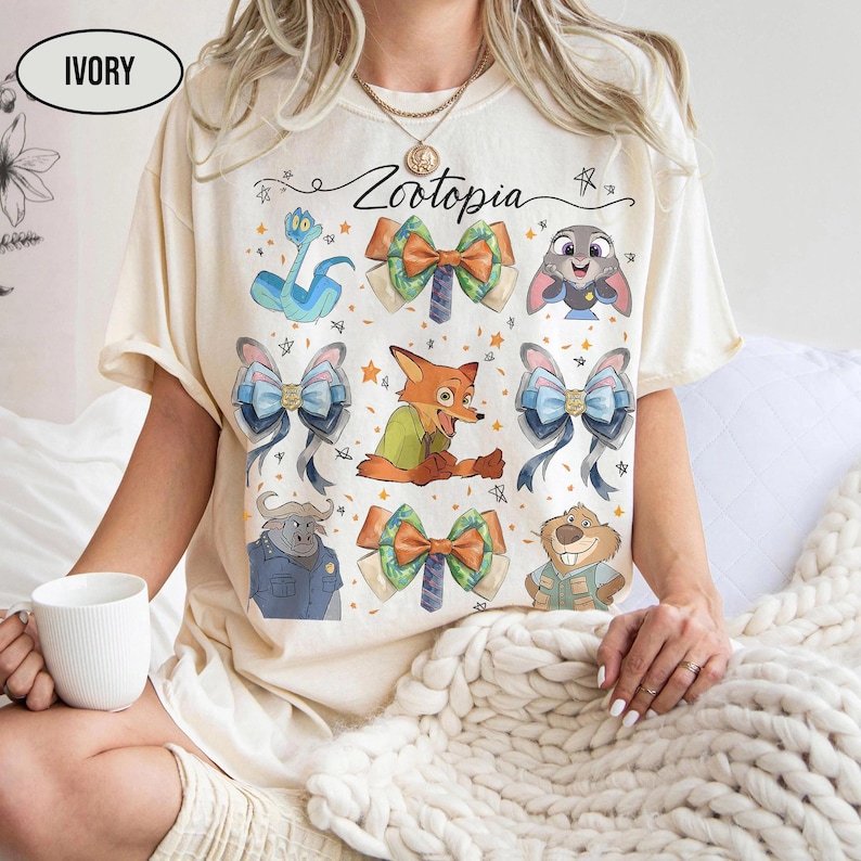 May include: An ivory t-shirt featuring the text Zootopia and a graphic print of various characters and bows. The design includes a blue snake, a fox, a buffalo, a beaver, a rabbit, and several colourful bows arranged around the central text.