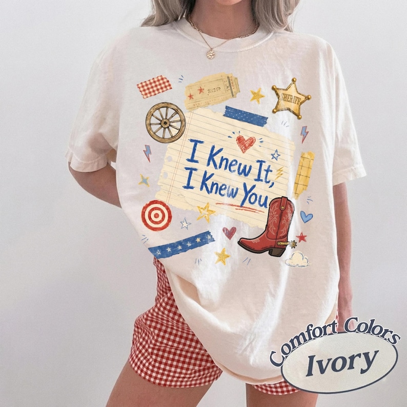 May include: An ivory t-shirt featuring a graphic print of a lined notebook paper with the text "I Knew It, I Knew You" in blue. The design includes a red cowboy boot, a gold sheriff star, a wagon wheel, a ticket, and various small colourful icons.