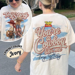 Może przedstawiać: Koszulka w kolorze kości słoniowej z nadrukiem graficznym zawierającym tekst Toy Story oraz You're a Cowboy like me Tay Story. Wz&oacute;r przedstawia stylizowaną ilustrację dw&oacute;ch postaci w kowbojskich kapeluszach, z kt&oacute;rych jedna gra na gitarze, na tle niebieskich pociągnięć pędzla.