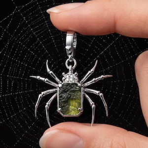 May include: A silver-toned spider pendant featuring a rectangular, textured olive-green stone as the body and a small cluster of clear stones on the head. The spider has eight slender, pointed legs and a metal lobster clasp attached to the top.
