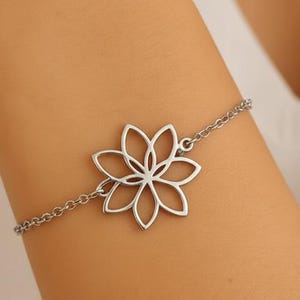 Peut inclure: Un bracelet argenté orné d'un motif floral ajouré. La fleur, à six pétales, est reliée à une fine chaîne. Le bracelet est porté au poignet, mettant en valeur son style élégant et minimaliste. La chaîne est faite de petits maillons.