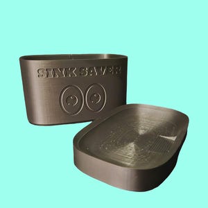 May include: A dark gray sink protector with the text "SINK SAVER" and two eye-shaped designs. The protector is rectangular with rounded edges, and a separate, smaller piece is next to it.