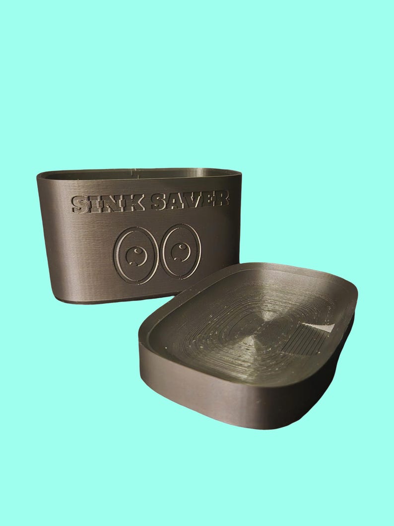 May include: A dark gray sink protector with the text "SINK SAVER" and two eye-shaped designs. The protector is rectangular with rounded edges, and a separate, smaller piece is next to it.
